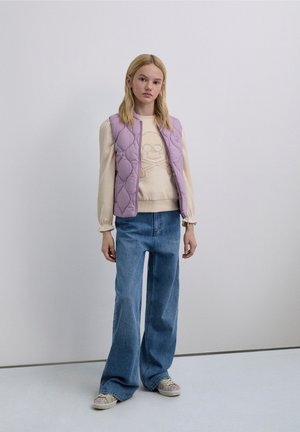 Gilet trapuntato lavanda sopra un maglione crema con grafica teschio, jeans a vita alta a gamba larga blu, e sneakers con motivo e suole chiare.