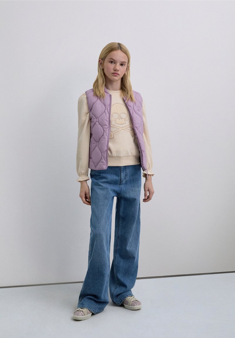 Veste matelassée lavande sur un pull crème avec motif crâne, jean large taille haute bleu, et baskets à motifs avec semelles claires.