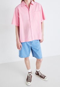 Kortärmad rosa skjorta med knappar och bröstficka, ihopparad med ljusblå denimshorts och bruna högstövlade sneakers.