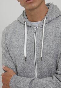 Siva zip-up hoodie izrađena od mekog materijala s teksturiranim završetkom. Ima kapuljaču, bijele vezice i cijelom dužinom zatvarač.