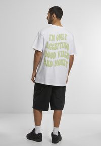 Camiseta de algodón blanca con corte oversize, con texto en verde lima en la parte posterior que dice: "SOLO ACEPTO BUENAS VIBRAS Y DINERO." Pantalones cortos negros y zapatillas.