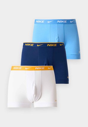 Homme debout pieds nus portant un boxer Nike bleu clair sur un fond blanc uni.