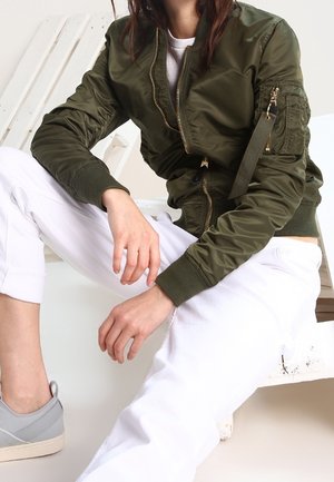 Personne portant une veste bomber vert olive, un pantalon blanc et des baskets grises, assise avec une jambe pliée sur un banc en bois blanc.