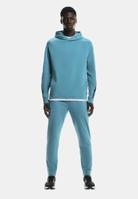 En mand står med ansigtet fremad iført en lyseblå hættejakke og matchende joggers samt sort-hvide sneakers på en hvid baggrund.