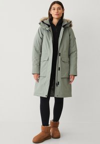 Lichtgroene parka jas met een gevoerde capuchon van bont, ritsen en drukknoopjes, voorzien van twee vo pockets en een trekkoord in de taille.