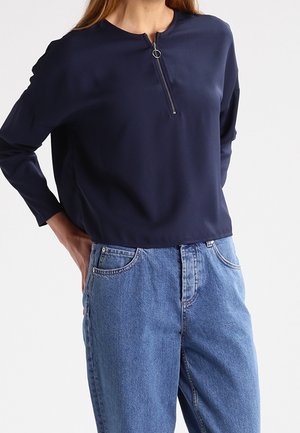 Person trägt ein marineblaues, langärmeliges Oberteil mit Vorderreißverschluss und weit geschnittene, hoch taillierte blaue Jeans, steht mit Händen in den hinteren Taschen.