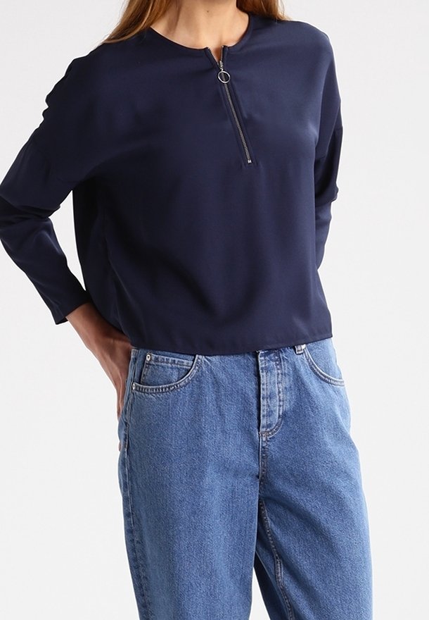 Personne portant un haut bleu marine à manches longues avec une fermeture éclair à l'avant et un jean bleu ample taille haute, debout les mains dans les poches arrière.