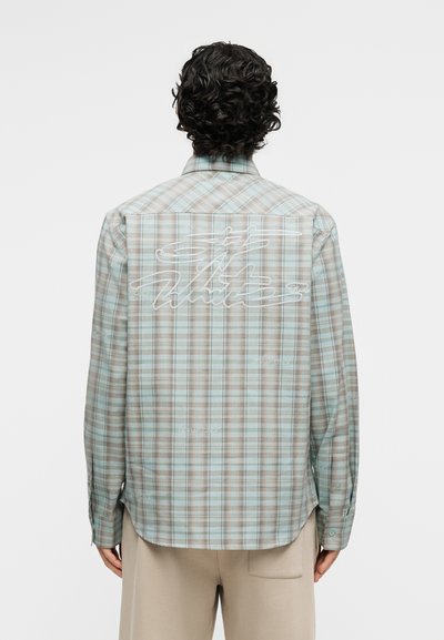 Άνδρας με σγουρά σκούρα μαλλιά φοράει ανοιχτό μπλε και μπεζ καρό πουκάμισο με λευκό γραφικό κείμενο στην πλάτη και μπεζ παντελόνι, στραμμένος με την πλάτη στην κάμερα.