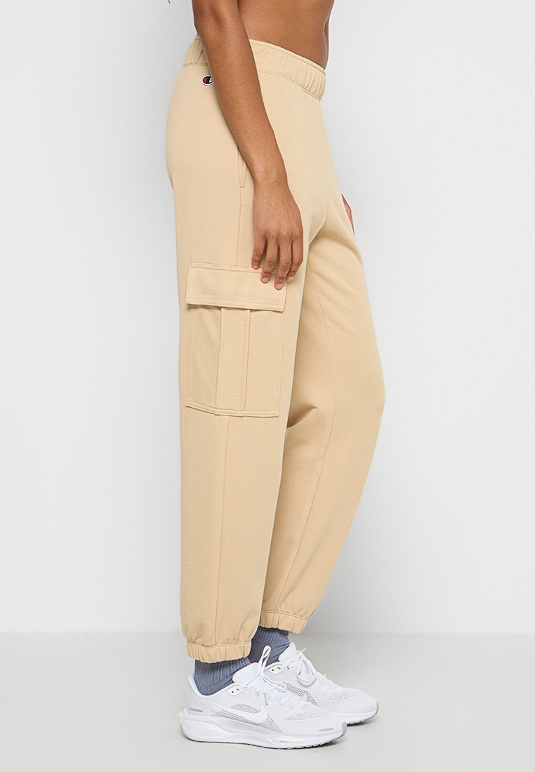 Pantalons de survêtement en coton beige avec des poignets élastiques et une poche cargo latérale. Dotés d'une taille à cordon de serrage. Assortis avec des baskets blanches et des chaussettes grises.