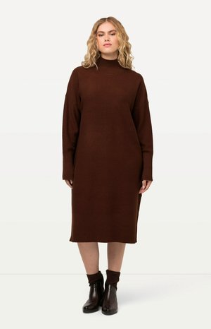 Bruine gebreide sweaterjurk met een hoge hals, lange mouwen en een losse pasvorm, voorzien van geribde boorden en zoom. Gepaarde met enkellaarsjes.