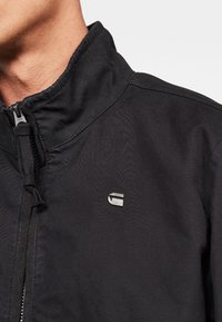 Man som bär en svart zip-up jacka med en liten metallisk logotyp på vänstra bröstet, kragen delvis dragkedjad.