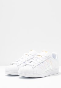 Bílé kožené tenisky s vroubkovanými špičkami, třemi iridiscentními pruhy a zlatými akcenty na jazyku. Hladká textura, lehký design.