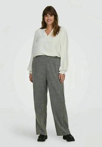 Donna sorridente che indossa una blusa bianca a maniche lunghe, pantaloni grigi a gamba larga e scarpe nere, in piedi davanti a uno sfondo bianco uniforme.