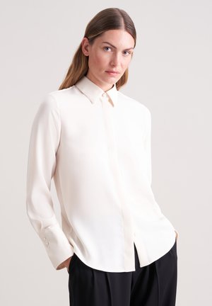 Blusa a maniche lunghe color crema realizzata in tessuto liscio, con colletto a punta e chiusura con bottoni. Presenta un orlo arrotondato e bottoni ai polsini.