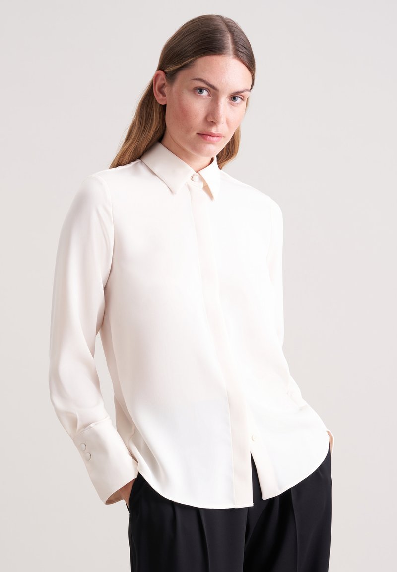 Long-sleeve, cream blouse made from smooth fabric with a pointed collar and button closure. Features rounded hem and cuff buttons.