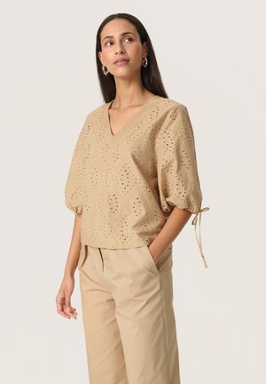 Beige cotton blouse with a V-neck, short puff sleeves, eyelet design, and adjustable ties at the sleeves. Paired with beige trousers.