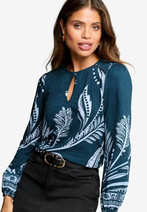 Türkisfarbige Bluse mit weißem botanischem Muster, runder Schlüssel-Ausschnitt, langen Puffärmeln und strukturiertem Finish, kombiniert mit einem schwarzen Gürtel und Rock.