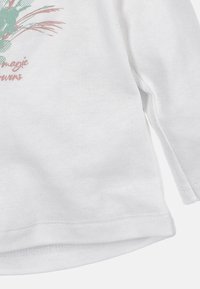 Biała koszula z długim rękawem z pastelową grafiką kaktusa i kwiatów oraz napisem „magic flowers” w stylu kursywy na wysokości klatki piersiowej.