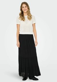Haut tricoté blanc à manches courtes avec un motif en dentelle, associé à une longue jupe noire à volants. Le modèle porte des bottines noires.