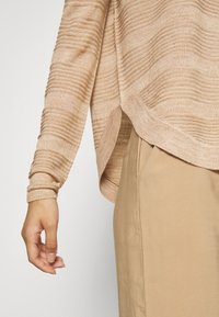 Bras gauche d'une personne portant un pull côtelé beige et un pantalon beige, avec la main détendue et les doigts légèrement recourbés.