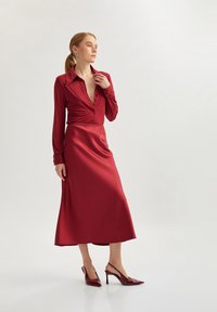 Vestido de seda rojo con mangas largas y un cuello, que cuenta con un cierre de botones en la parte delantera y una falda fluida. Combinado con unos zapatos de tacón burgundy.