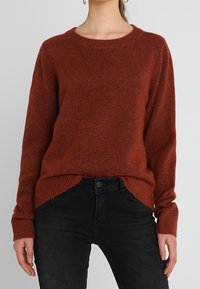 Rostfarbener Strickpullover mit Rundhalsausschnitt, langen Ärmeln und seitlichen Schlitzen, mit einer weichen Textur sowie gerippten Bündchen und Saum.