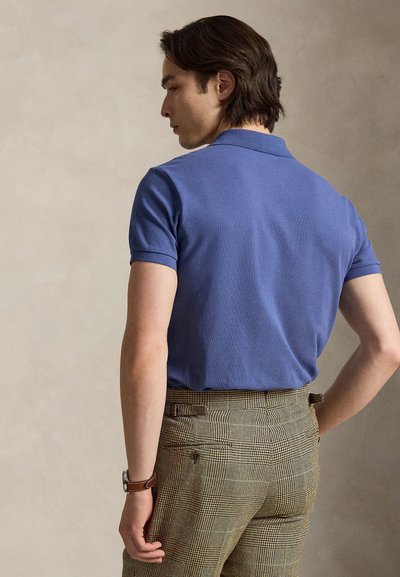 Mann som vender ryggen til, iført en blå kortermet polo og brune rutete bukser med en lærarmbåndsur.