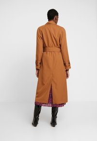 Abrigo largo marrón tipo gabardina con cinturón, cuello alto y puños abotonados. Cuenta con una abertura central en la parte trasera, usado sobre un vestido estampado.