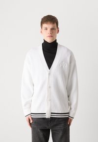 Cárdigan de punto blanco con escote en V profundo, seis botones y puños y dobladillo de canalé negro. Presenta un patrón texturizado en la parte superior del cuerpo.