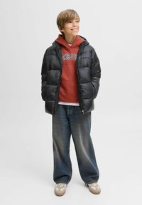 Doudoune noire avec capuche, sweat à capuche rouge en dessous, jean bleu ample et baskets beiges avec accents blancs.