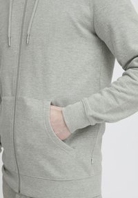 Felpa grigia con zip realizzata in tessuto morbido, dotata di tasche laterali, polsini a coste e cappuccio con cordino. Texture liscia con dettagli delle cuciture.
