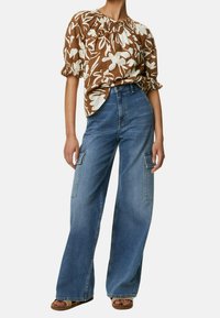 Blouse fleurie marron avec manches courtes bouffantes, associée à un pantalon cargo en jean bleu taille haute à jambes larges, doté de grandes poches latérales.