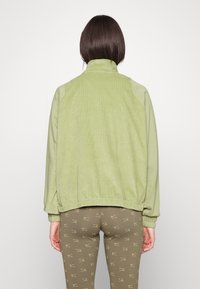 Lys grøn jakke med en kord-tekstur, lynlås krave og elastisk kant, parret med brune leggings med et gentaget logo mønster.
