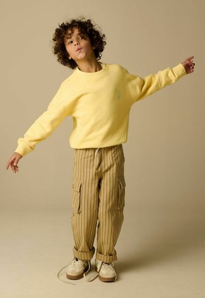 Enfant aux cheveux bouclés portant un sweat-shirt jaune et un pantalon cargo rayé, s'équilibrant les bras étendus sur un fond beige.