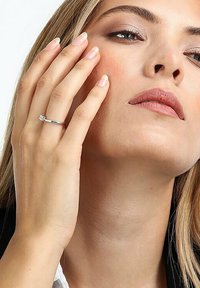 Silberner Ring mit einem kleinen, klaren Edelstein, der an einer Hand getragen wird, die das Gesicht berührt. Der Finger hat eine sanfte, natürliche Maniküre.