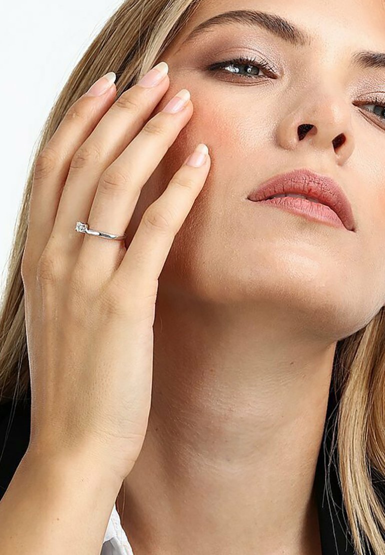 Silberner Ring mit einem kleinen, klaren Edelstein, der an einer Hand getragen wird, die das Gesicht berührt. Der Finger hat eine sanfte, natürliche Maniküre.