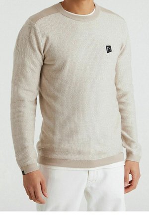 Homme portant un pull beige texturé à col rond avec un petit logo noir sur la poitrine, associé à un pantalon blanc.
