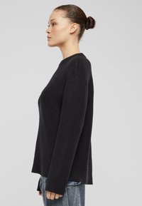 Schwarzer Strickpullover mit geribbelter Textur, Rundhalsausschnitt und langen Ärmeln, mit Schlitzen am Saum, kombiniert mit ausgewaschenen Jeans.