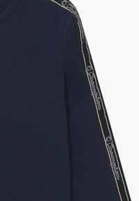 Marinblå ärm med svart rand som har den upprepade vita Champion-logotypen i kursiv som löper längs armen.