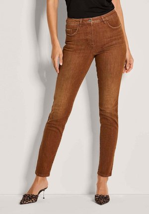 Frau in Slim-Fit braunen Jeans und High-Heel-Mules mit Leopardenmuster, die entspannt mit den Händen an einer hellen Wand steht.