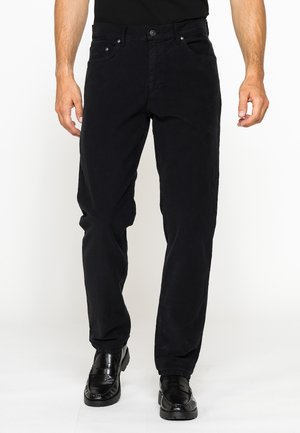 Uomo che indossa pantaloni neri a gamba dritta, mocassini neri e una camicia nera, in piedi davanti a uno sfondo bianco.