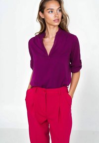 Fioletowa bluzka z dekoltem w serek i długim rękawem połączona z jasnoróżowymi spodniami o dopasowanym kroju. Bluzka ma subtelne fałdy, a spodnie posiadają kieszenie po bokach.