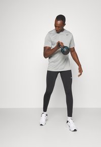 Mann i grå Nike t-skjorte med kort erme, svarte leggings, holder en svart kettlebell med blå detaljer, iført hvite joggesko.