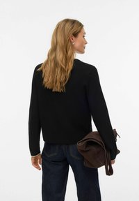 Črni pleteni pulover sproščenega kroja z okroglim izrezom in dolgimi rokavi, kombiniran z visokim pasom temno modrih kavbojk in rjavo torbo.