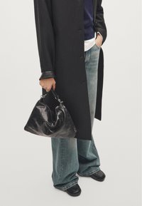 Sac en cuir noir au design souple et décontracté, avec des détails en métal et une courte anse, porté à côté d'un jean large bleu clair.