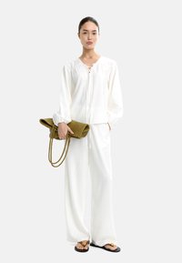 Biała, lekka bluzka z przodem na sznurki, zestawiona z luźnymi białymi spodniami. Trzyma teksturowaną oliwkowo-zieloną torebkę kopertówkę z plecionymi uchwytami.