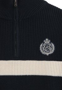 Marinblå stickad tröja med en halvdragkedja vid kragen, har en krämfärgad horisontell rand och en broderad logotypmärke på bröstet.