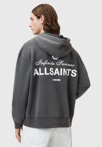 Szary, oversize'owy hoodie z kapturem na sznurku, z białym nadrukowanym tekstem na plecach oraz prążkowanymi mankietami i dołem. Miękka tekstura materiału.