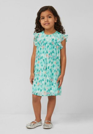 Junges Mädchen mit lockigem Haar, das ein türkisfarbenes, geblümtes, plissiertes Kleid und silberne Mary-Jane-Schuhe trägt, steht vor einem einfarbigen Hintergrund.