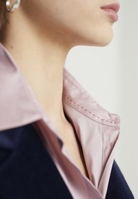 Indumento con colletto in raso rosa chiaro con cuciture a contrasto, abbinato a un corpo in tessuto scuro e strutturato. Vista ravvicinata.
